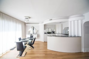 A modern kitchen with a curved island, a dining area with a glass table and chairs, and a staircase leading to an upper level. - Water Street Retreat 09 (Wilmington)