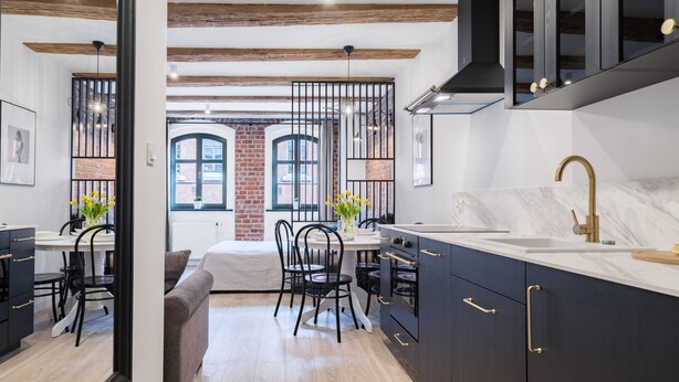 A modern kitchen with dark cabinetry, a marble countertop, and a gold faucet. There is a dining area with a table and chairs, and a view of a brick building through a window. - Miodowa Faktoria 7 (Torun)