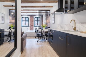 A modern kitchen with dark cabinetry, a marble countertop, and a gold faucet. There is a dining area with a table and chairs, and a view of a brick building through a window. - Miodowa Faktoria 7 (Torun)