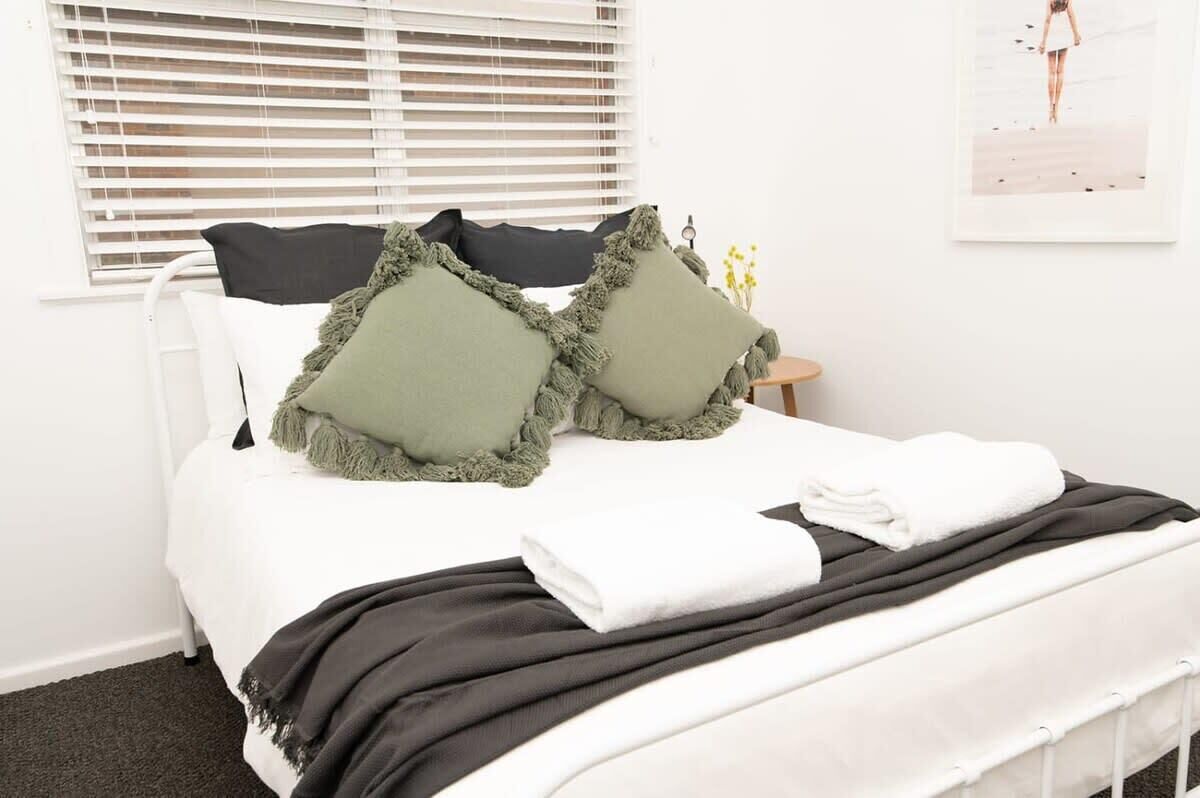 Ein ordentlich bezogenes Bett mit weißer Bettwäsche, zwei grünen Kissen und einer schwarzen Tagesdecke.