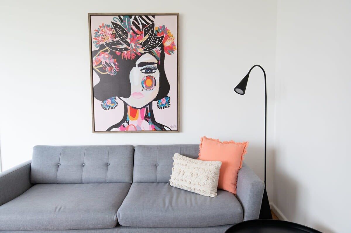 Ein graues Sofa mit einem weiß-orangefarbenen Kissen, eine schwarze Stehlampe und ein gerahmtes abstraktes Gemälde an der Wand.
