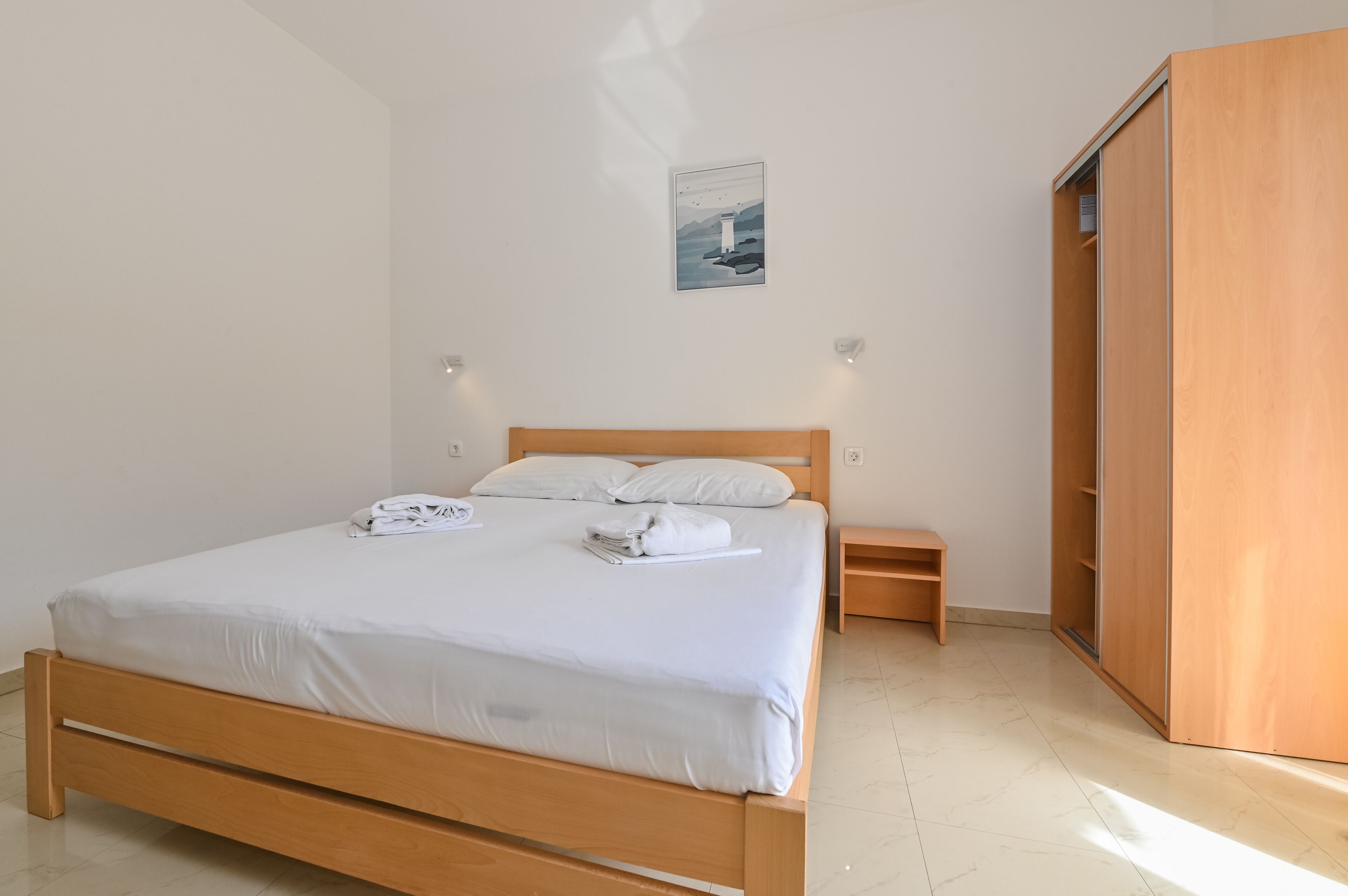 Una cama bien hecha con ropa blanca, una mesita de noche, un armario de madera y un aplique en la pared.