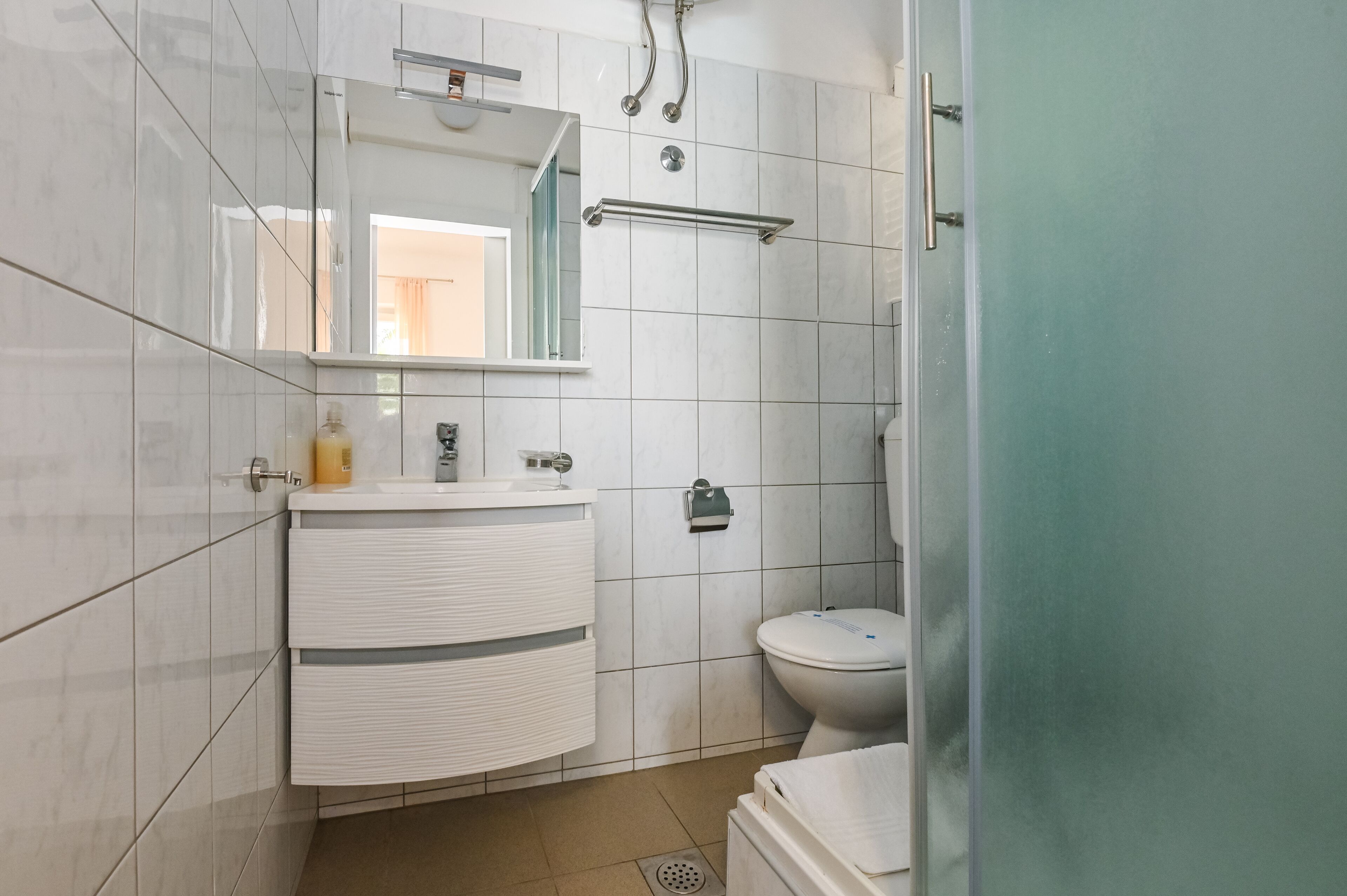 Casa de banho moderna com azulejos brancos, lavatório branco com espelho, sanita e zona de duche.