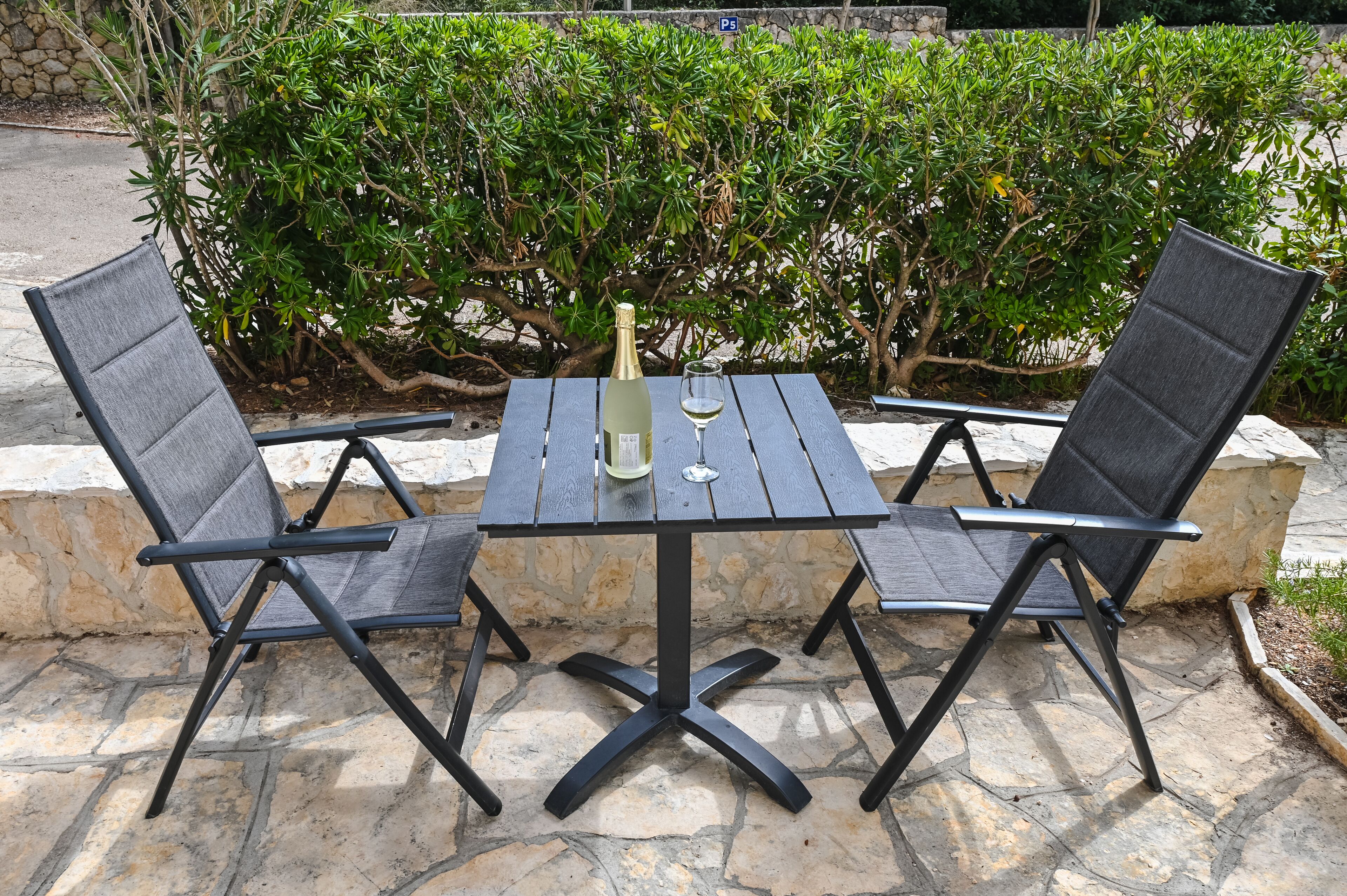 A table and two chairs with a bottle and glass on it, set on a stone patio with a hedge in the background.