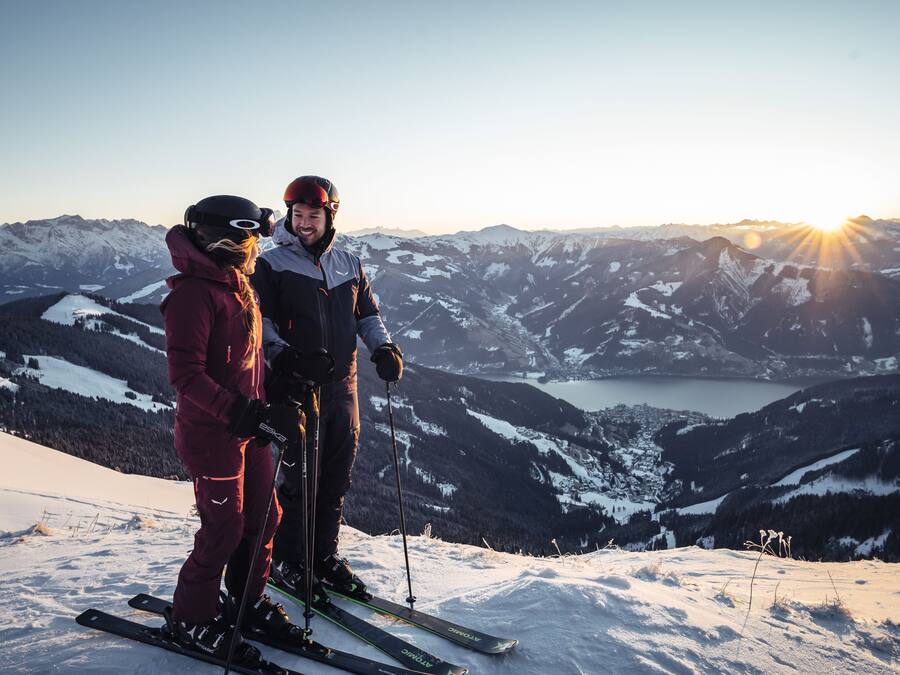 A couple on skis at a mountain peak with a view of snow-covered mountains and a lake.