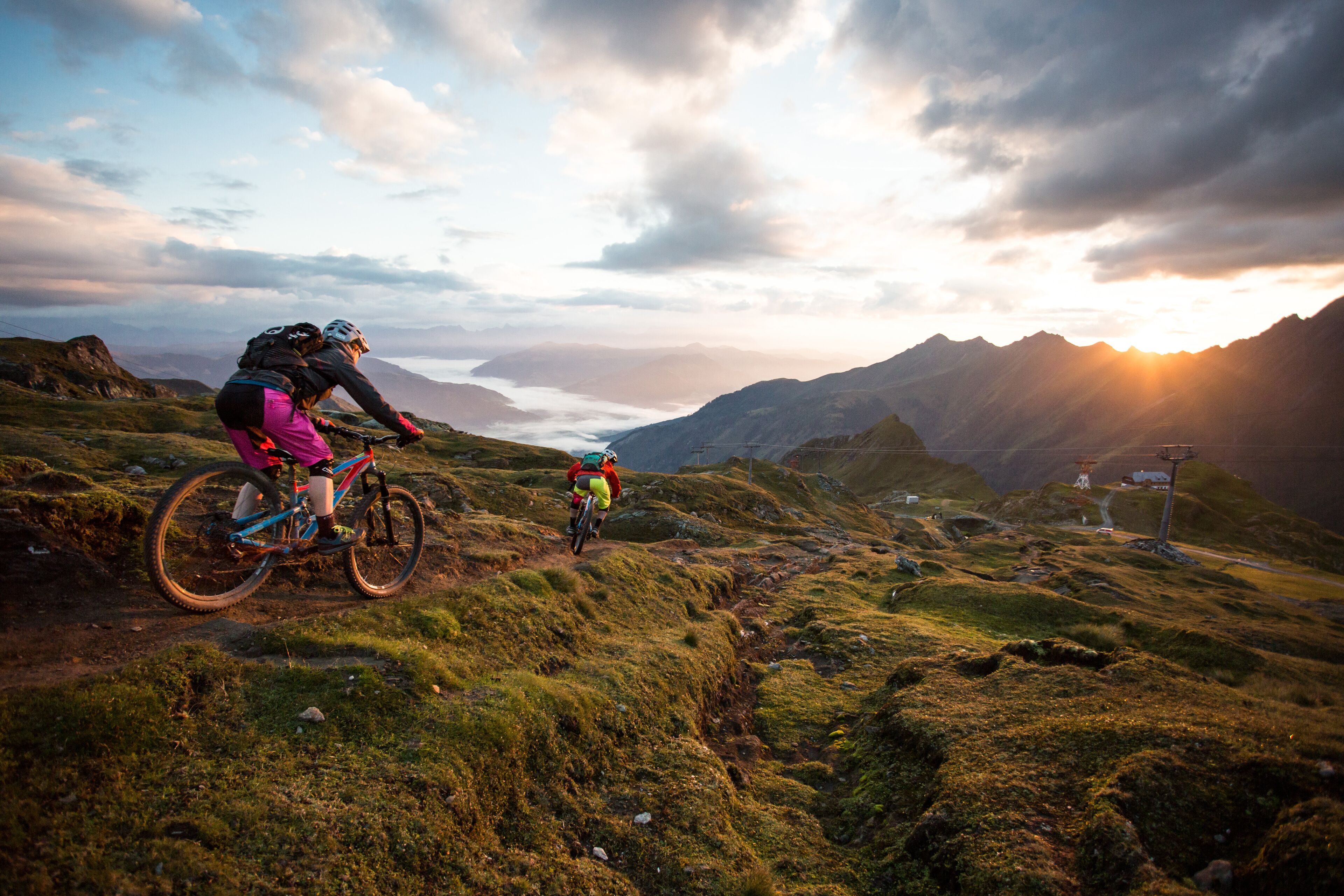 En mountainbikeled med två cyklister, omgiven av frodig grönska och berg i bakgrunden.