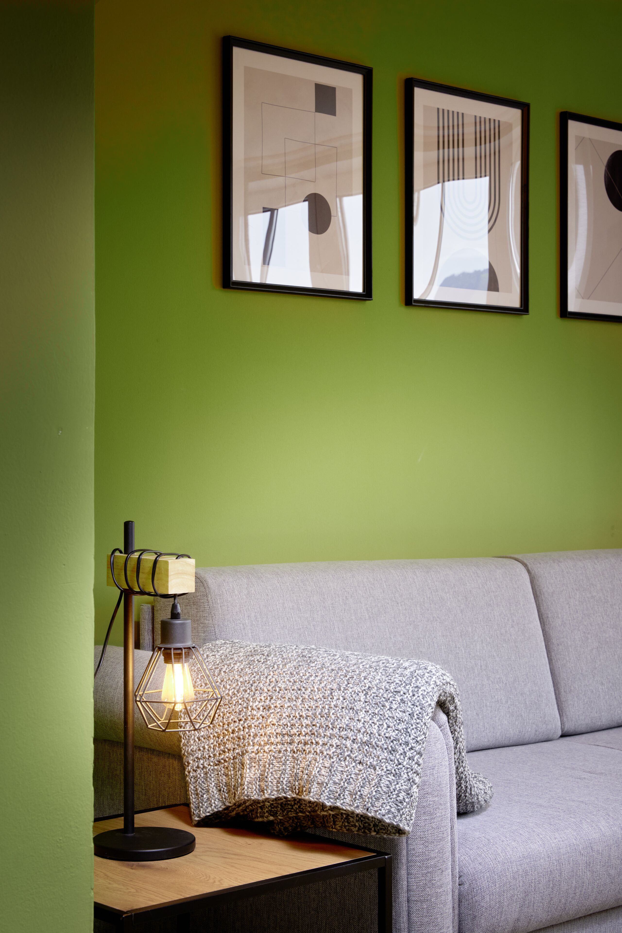 Ein Zimmer mit grüner Wand, einem grauen Sofa und einem Tisch mit Lampe. Drei gerahmte Bilder hängen an der Wand.