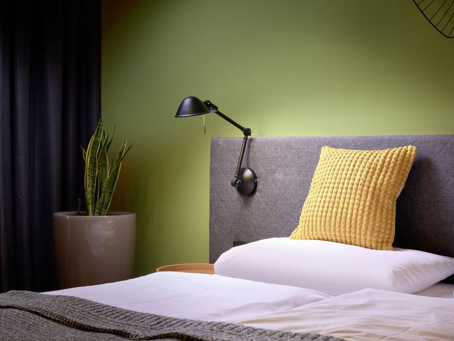 A neatly made bed with white linens, a grey headboard, and a yellow textured pillow. A black desk lamp is on the bedside table, and a potted plant is visible in the corner.