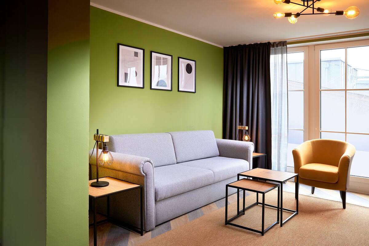 A modern living room with a grey sofa, a yellow armchair, and a small wooden table. There are three framed pictures on the wall and a floor lamp.