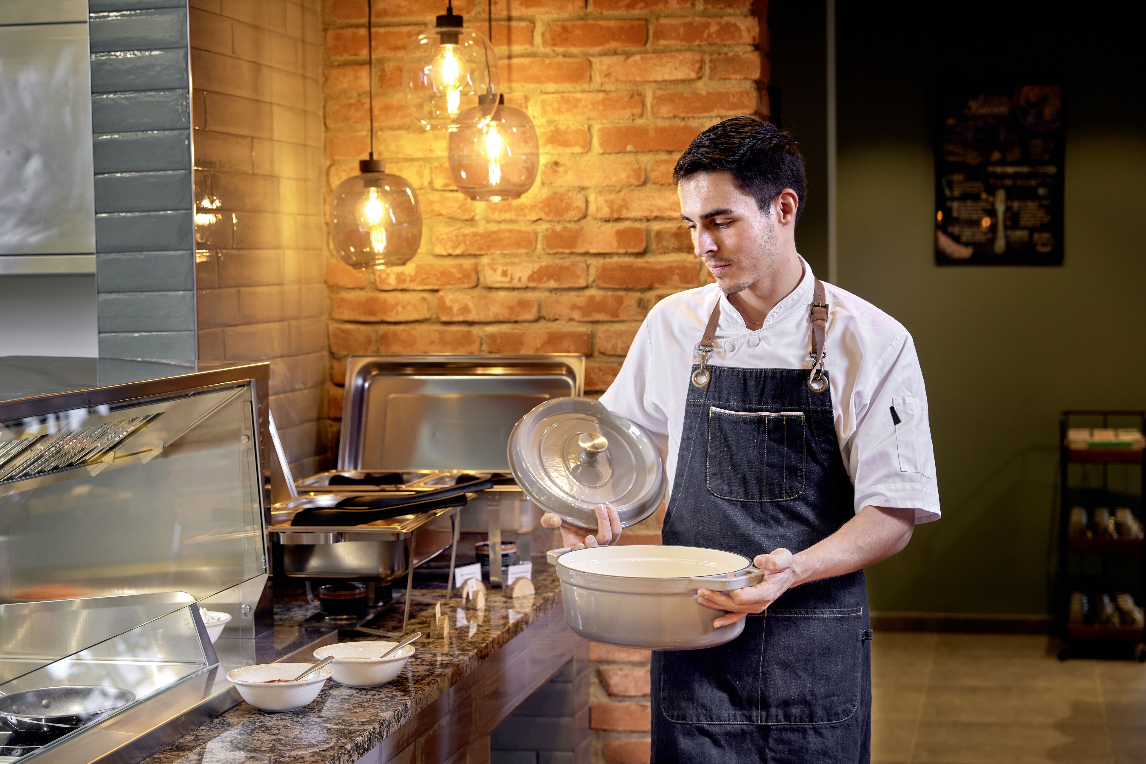 A buffet setup with a chef holding a pot, a glass display case, and a brick wall with hanging lights.