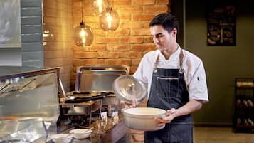 Montaje de bufé con un cocinero sosteniendo una olla, una vitrina de cristal y una pared de ladrillo con luces colgantes.