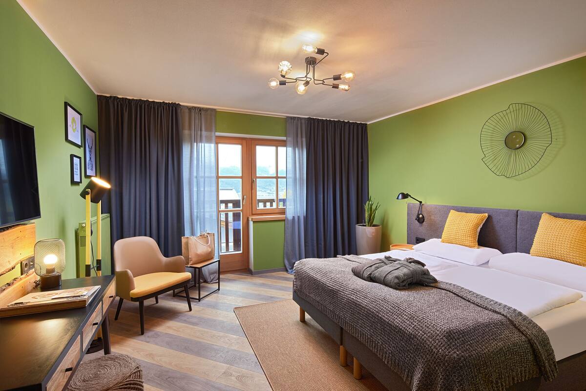 A modern hotel room with a large bed, a desk with a lamp, a chair, and a view through a window with curtains.