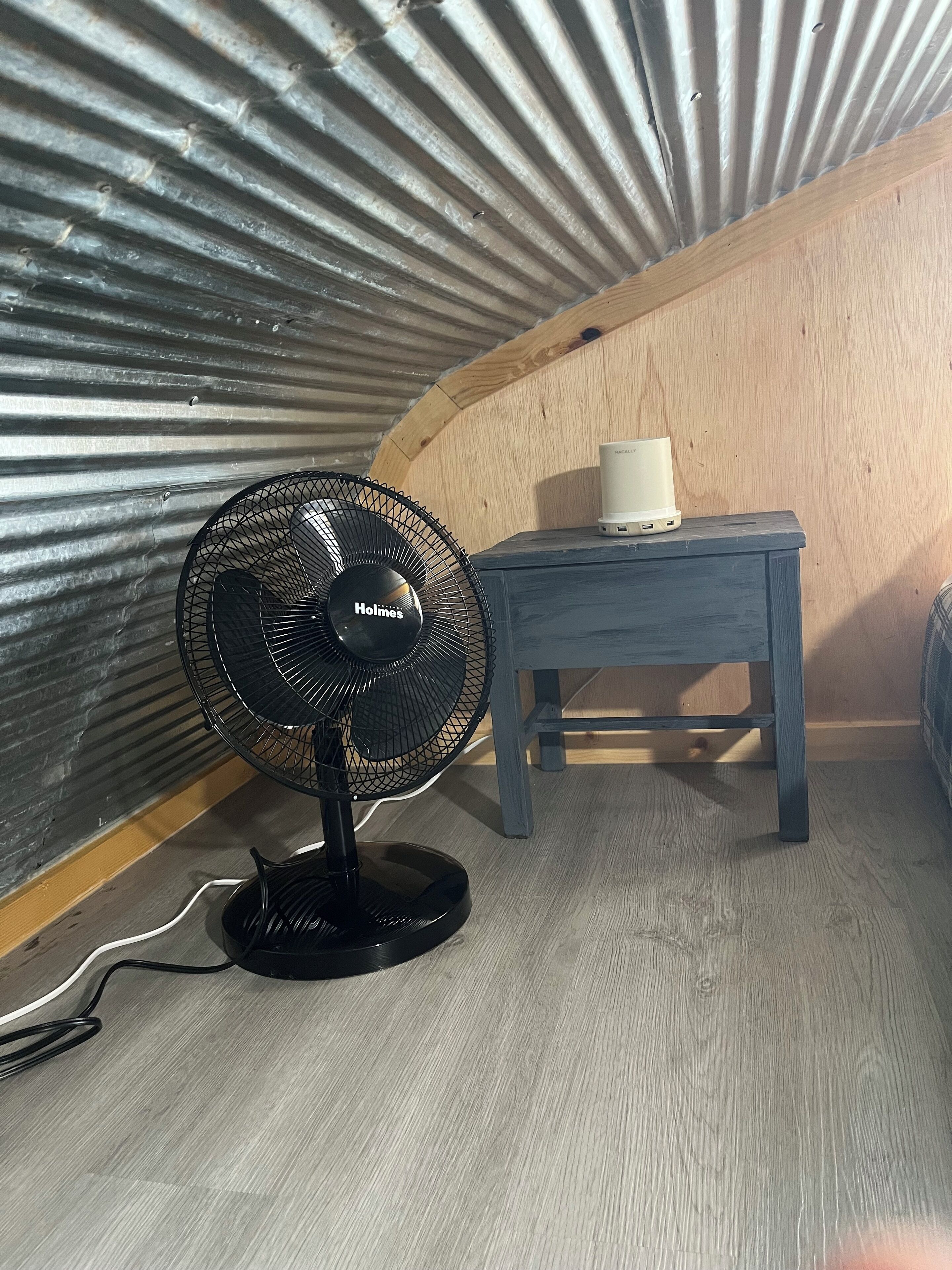 Una habitació petita amb un ventilador Holmes negre, una taula de fusta amb un dispositiu blanc i una paret de fusta.