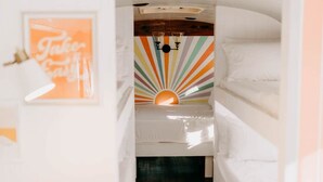 A narrow hallway with a wooden floor leading to a room with a colorful sunburst wall design and a bed with white bedding. - Lost Hill Lake Sunshine Skoolie, The Sunshine Skoolie - Lost Hill Lake (Saint Clair)