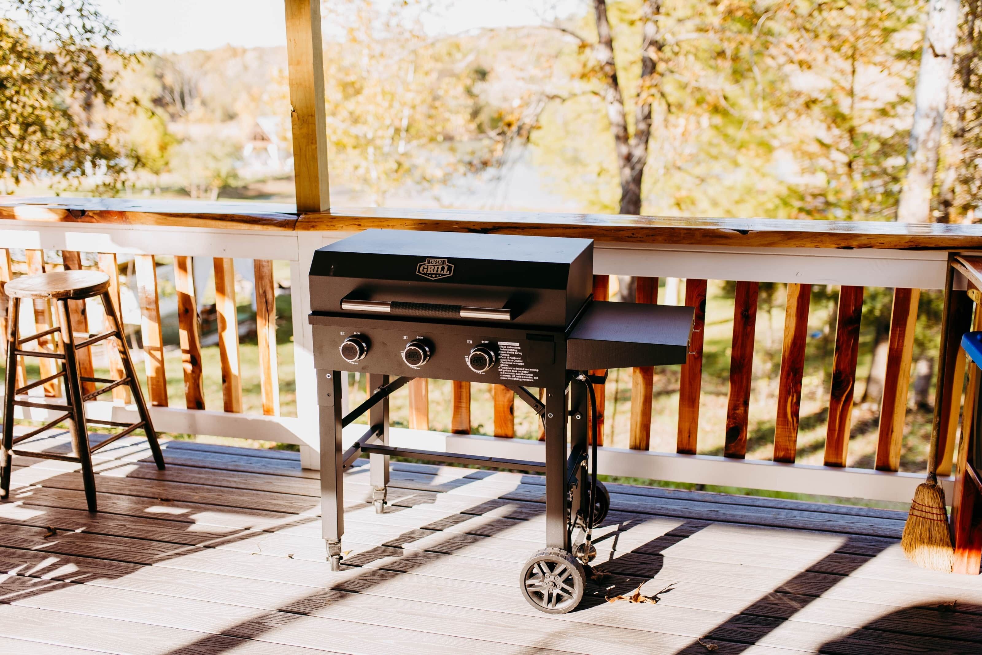 Una parrilla para barbacoa en una terraza de madera con barandilla, un taburete de madera y una escoba apoyada en la barandilla.