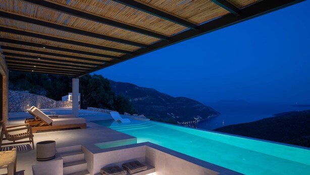A luxurious outdoor infinity pool area with a view of the sea and mountains, wooden lounge chairs, and a stone wall. - Armante Supreme Villas-Villa Nicole (Fterno)