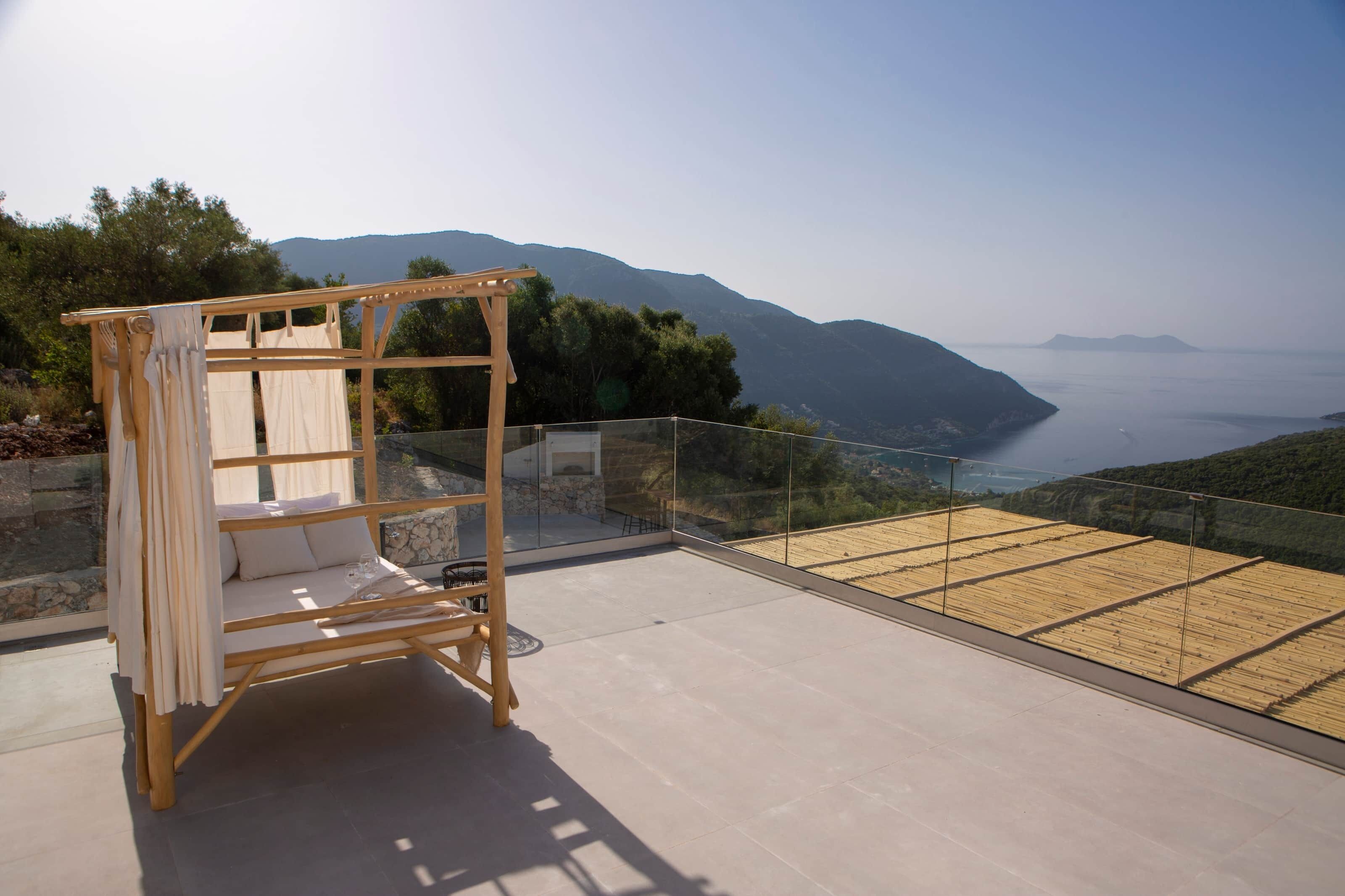 Ein Bett aus Holz mit weißen Laken auf einem Balkon mit Glastrennwand, mit Blick auf eine Küstenlandschaft mit Bergen und dem Meer.