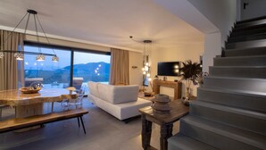 A modern living room with a large window, a sofa, a dining table, and a staircase. - Armante Supreme Villas-Villa Nicole (Fterno)