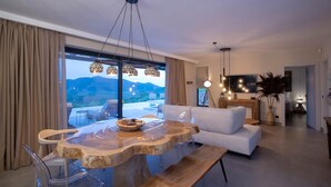 A modern living room with a large wooden dining table, white sofa, and a view of mountains through large windows. - Armante Supreme Villas-Villa Nicole (Fterno)