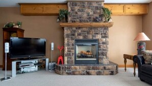 A living room with a stone fireplace, a flat-screen TV, and a wooden entertainment unit. - Big White Beauty! Sleeps 10.  Ski in/Ski out Chalet with Private Hot Tub. (Beaverdell)