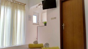 A hotel room with a bed, a TV mounted on the wall, a wooden door, and a window with curtains. - Studio for 2 people in a quiet part of town (Baška)