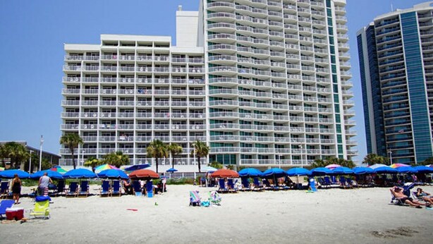 Een groot hotel met meerdere balkons, een strandgedeelte met parasols en een zandstrand.