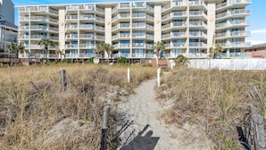 A beachfront hotel with multiple balconies, a sandy path leading to the entrance, and a white fence. - Crescent Sands of Windy Hill  F4-3BR/3BA Oceanfront condo in Crescent Beach (North Myrtle Beach)
