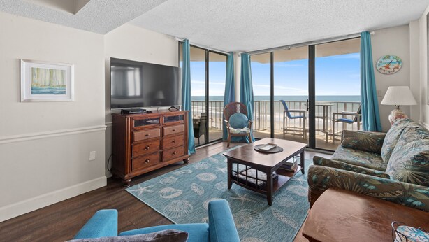 A living room with a large TV, wooden furniture, a patterned rug, and a view of the ocean through large windows. - Crescent Sands of Windy Hill  F4-3BR/3BA Oceanfront condo in Crescent Beach (North Myrtle Beach)