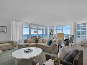 A spacious living room with a large window overlooking the sea, a beige sofa, a white coffee table, and a dining area with a chandelier.