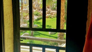 A view through a window with metal bars, featuring a grassy area with palm trees and a building in the background. - Top Floor Large 2 Bedroom  Ocean/Beach Views Vaulted Ceiling Lots of Windows (Cabo San Lucas)