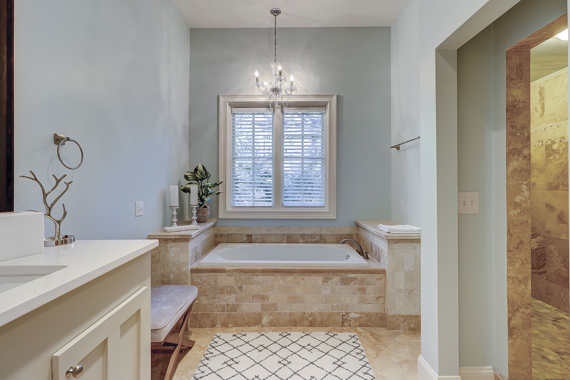 Une salle de bain avec une baignoire, une fenêtre avec des stores, un lustre et une plante.