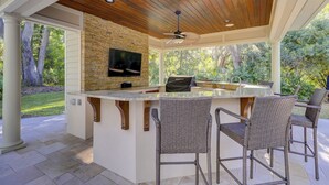 Una cocina al aire libre techada con pared de piedra, un televisor y sillas de bar de mimbre.
