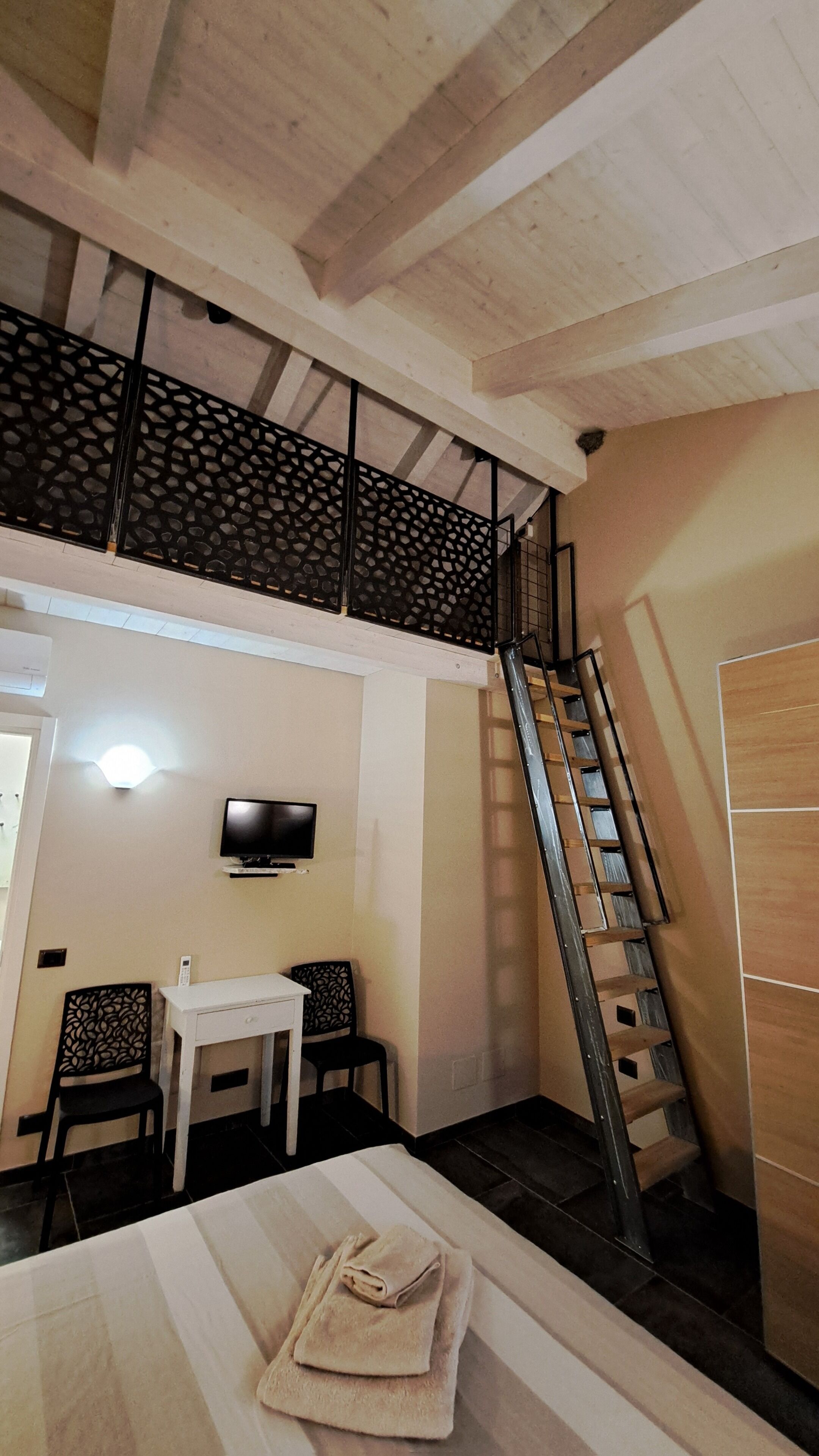 Habitació amb escriptori blanc, dues cadires, televisió penjada a la paret, escala i porta de fusta.