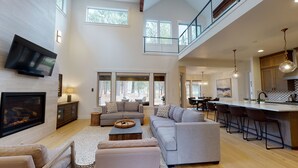 A spacious living area with a large sofa, a coffee table, and a TV mounted on the wall. There is a balcony with a railing and a kitchen area with bar stools and a hanging light fixture. - Caldera Getaway with Access to Forest House (Sunriver)