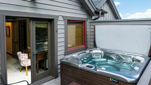 A hot tub with a control panel, a wooden deck, a chair, and a door leading to an interior space.