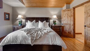 A bedroom with a wooden ceiling, a large bed with white bedding, two bedside tables with lamps, and a wooden door.