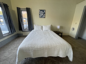 Une chambre à coucher avec un grand lit, une fenêtre avec des rideaux, une table de chevet avec une lampe et une œuvre d’art accrochée au mur.