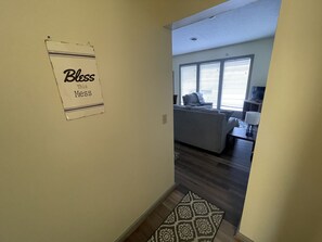 A hallway leading to a living room with a gray sofa, a patterned rug, and a sign that reads "Bless This Mess".