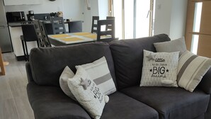 Una sala de estar moderna con un sofá gris oscuro, una zona de comedor con una mesa amarilla y una cocina al fondo.