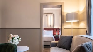 A hotel room with a green chair, a round table, a sofa, a lamp, and a bed with a headboard.