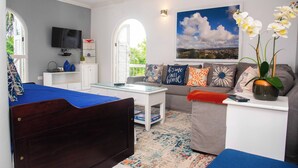 A modern living room with a bed, a sofa, a coffee table, and a TV. There is a window with a view of the outdoors.