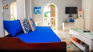 A bedroom with a blue bed, white walls, a TV, and a view of the outdoors.