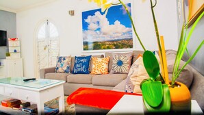 A living room with a sofa, a glass coffee table, a red ottoman, and a vase with orchids. There is a painting of a cityscape on the wall.