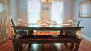 Comedor con mesa de madera oscura, dos sillas, un banco, una araña y una ventana con la palabra "Lake" en la pared.