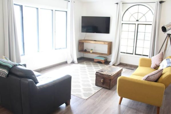 A living room with a black sofa, a yellow armchair, a wooden coffee table, and a TV on the wall.