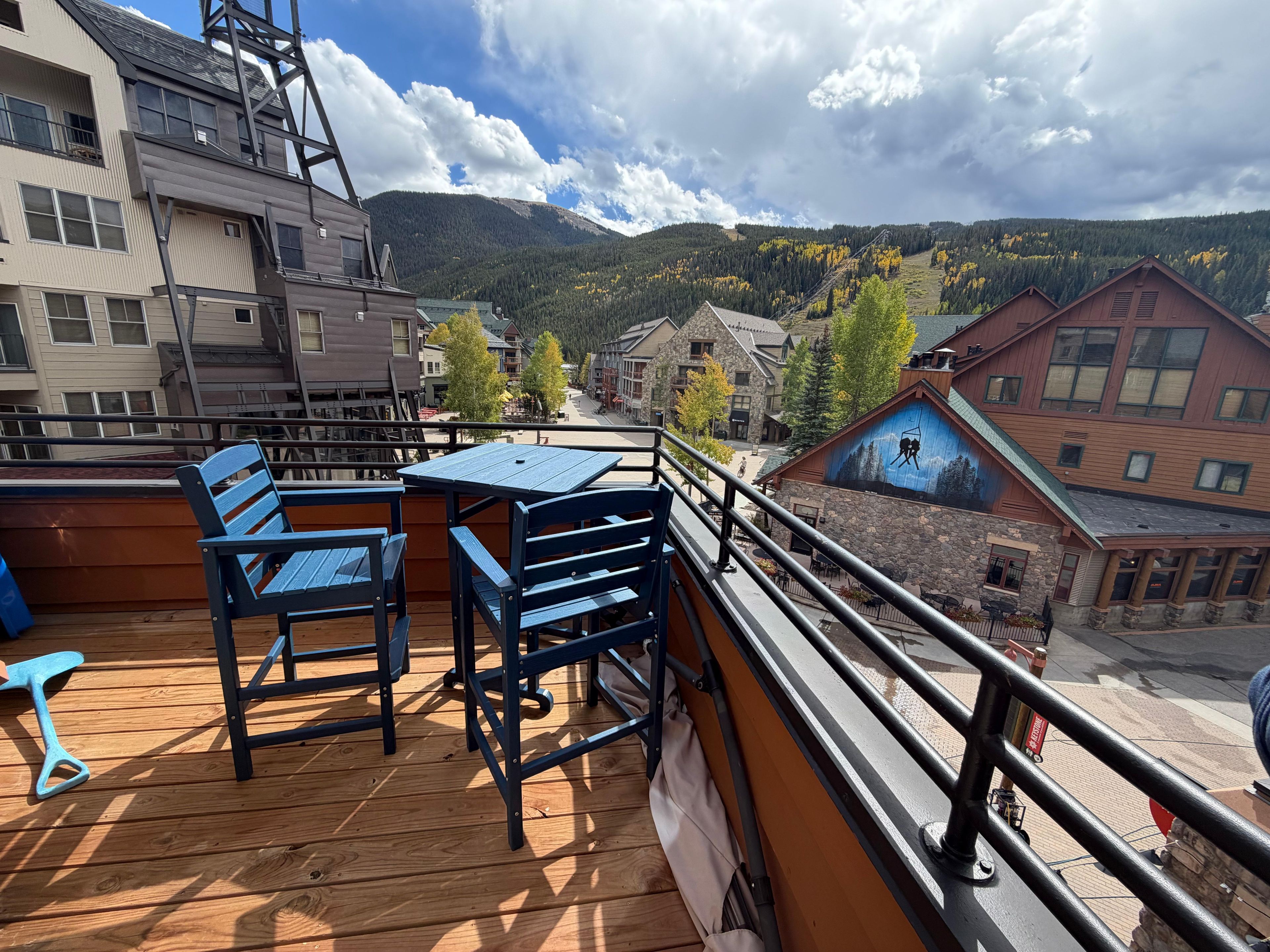 A wooden deck with a table and chairs, overlooking a town with buildings and a mountain in the background.