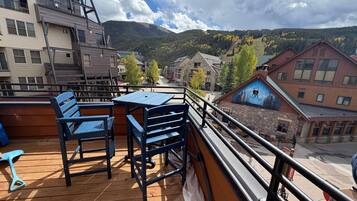 A wooden deck with a table and chairs, overlooking a town with buildings and a mountain in the background.
