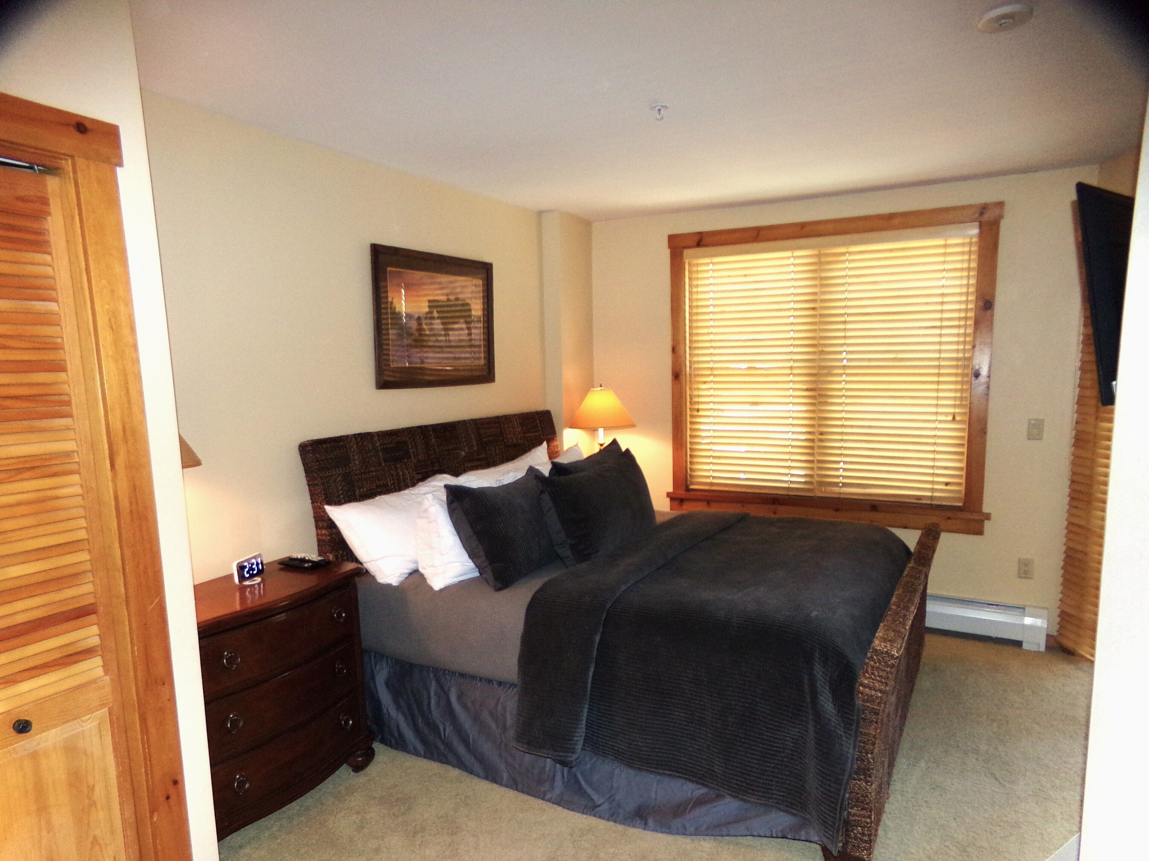 A bedroom with a bed, a nightstand, a lamp, a window with blinds, and a framed picture on the wall.