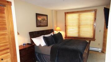A bedroom with a bed, a nightstand, a lamp, a window with blinds, and a framed picture on the wall.