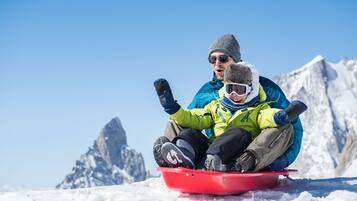 A person sitting on a red sled on a snowy surface with a mountainous background.